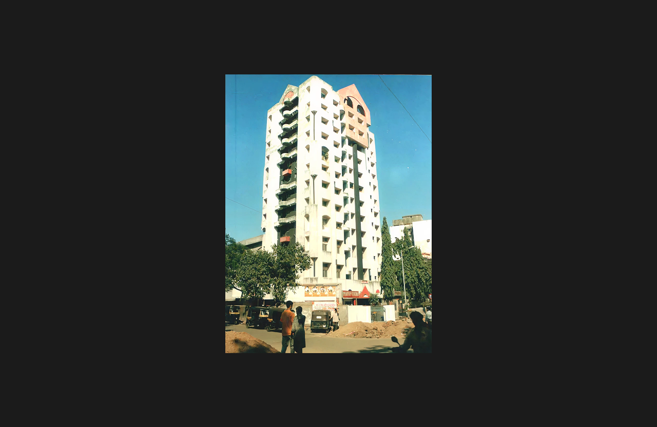 A tall residential building with a white façade and geometric window recesses, featuring external balconies on one side and a distinctive pink architectural crown at the top, photographed from a street-level perspective with pedestrians, trees, and vehicles in the foreground under a clear blue sky.