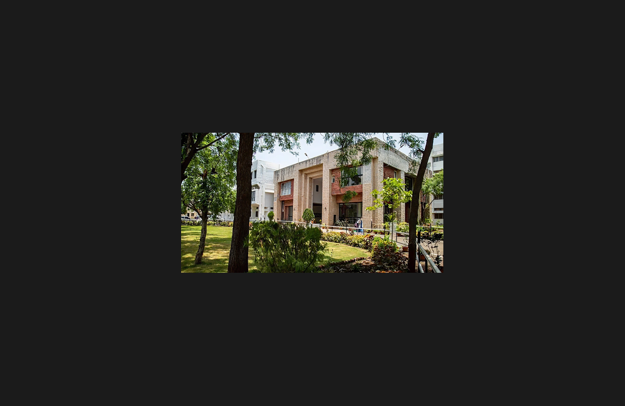 A modern institutional building with a contemporary architectural façade featuring stone cladding and large glass windows, surrounded by landscaped gardens, trees, and a pedestrian walkway.