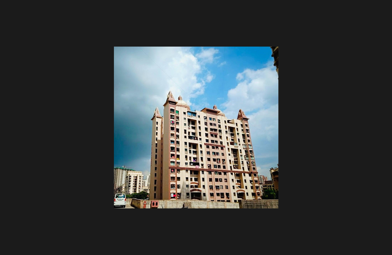 A wide-angle view of a multi-wing high-rise residential apartment building with decorative architectural turrets and beige exterior finishes, captured under a bright blue sky with dramatic clouds, with surrounding urban buildings and vehicles in the foreground.