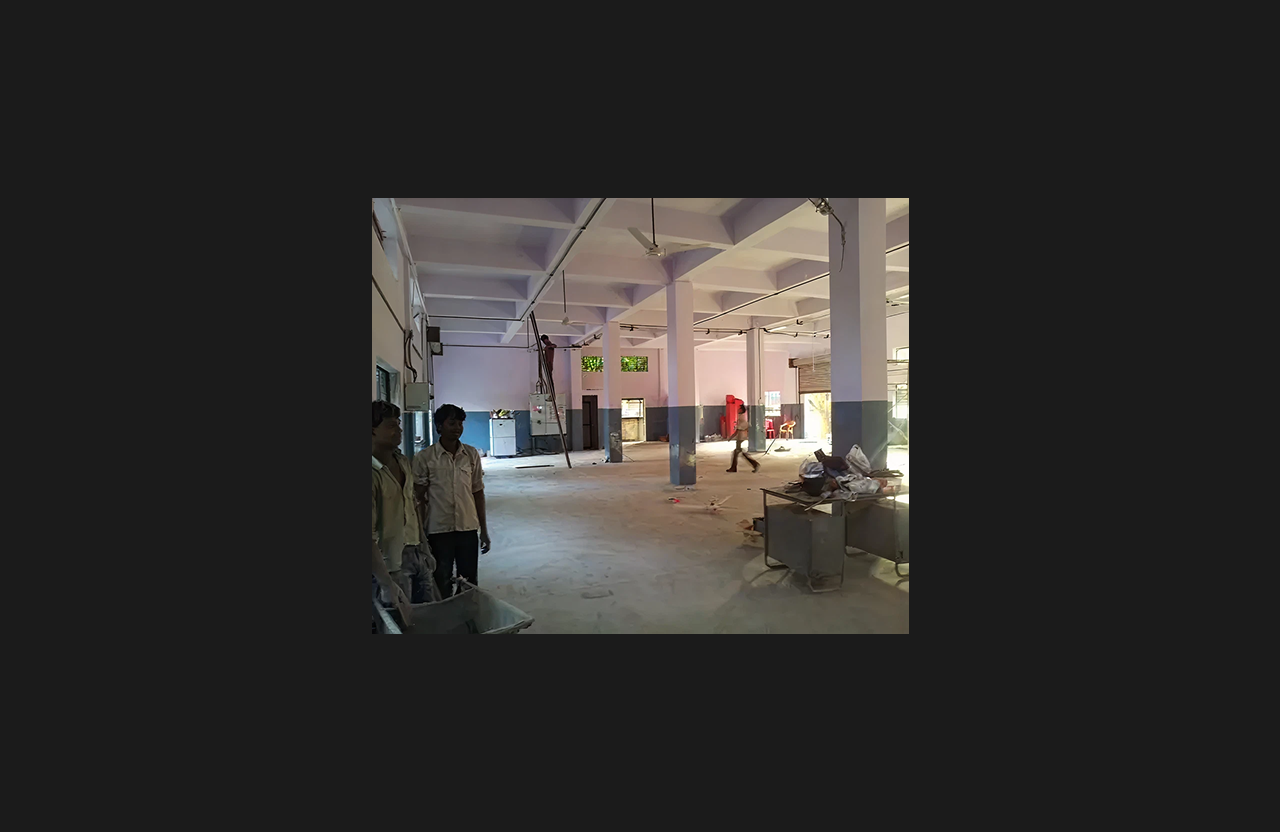 Construction progress inside a spacious hall with workers, ladders, and equipment visible, unfinished flooring, exposed ceiling framework, and open shuttered entry areas providing daylight.
