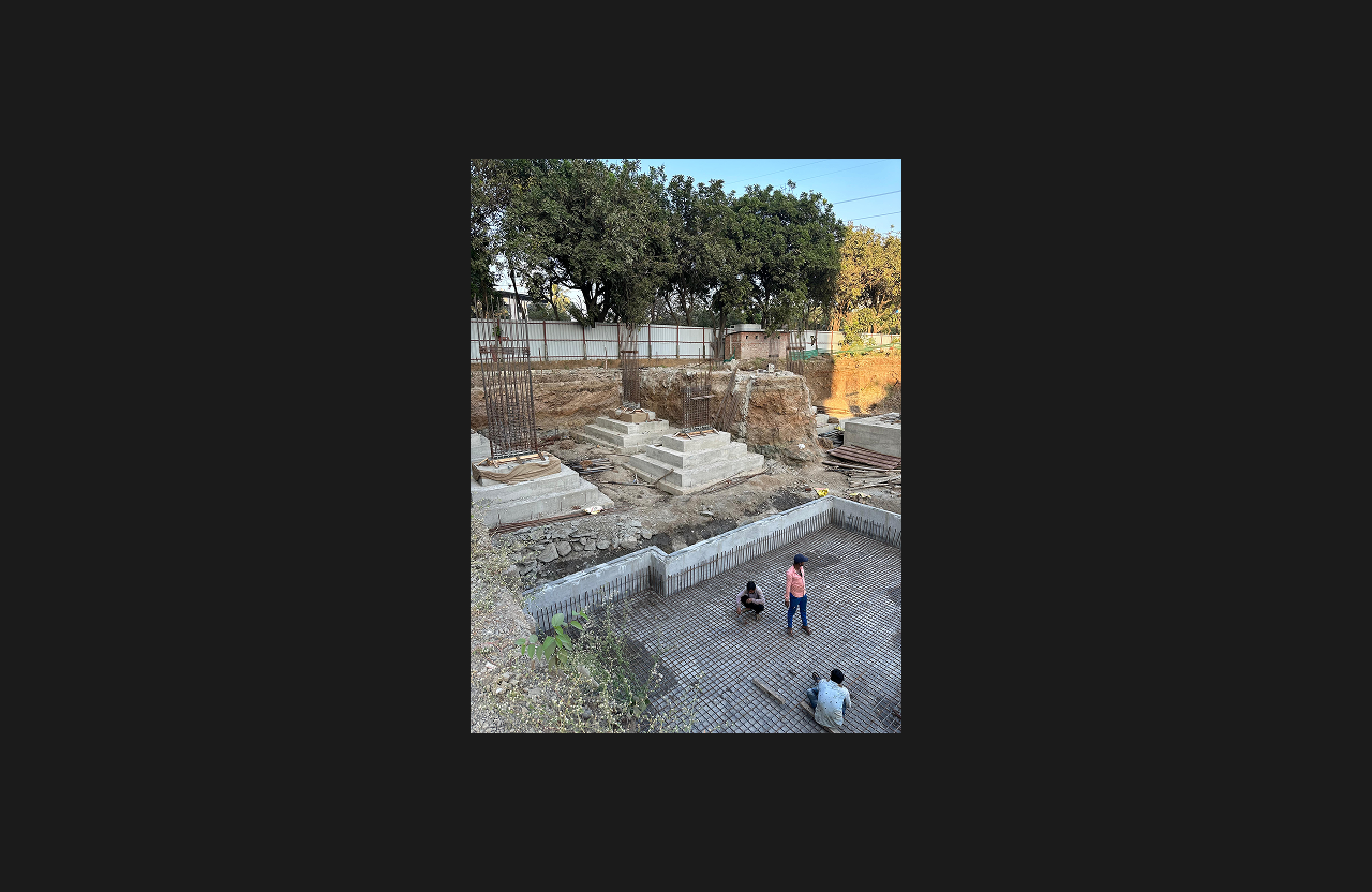A construction site showing early-stage foundation work with reinforced steel grid and concrete footing structures, and three workers inspecting progress inside the excavated area, with trees and fencing in the background.