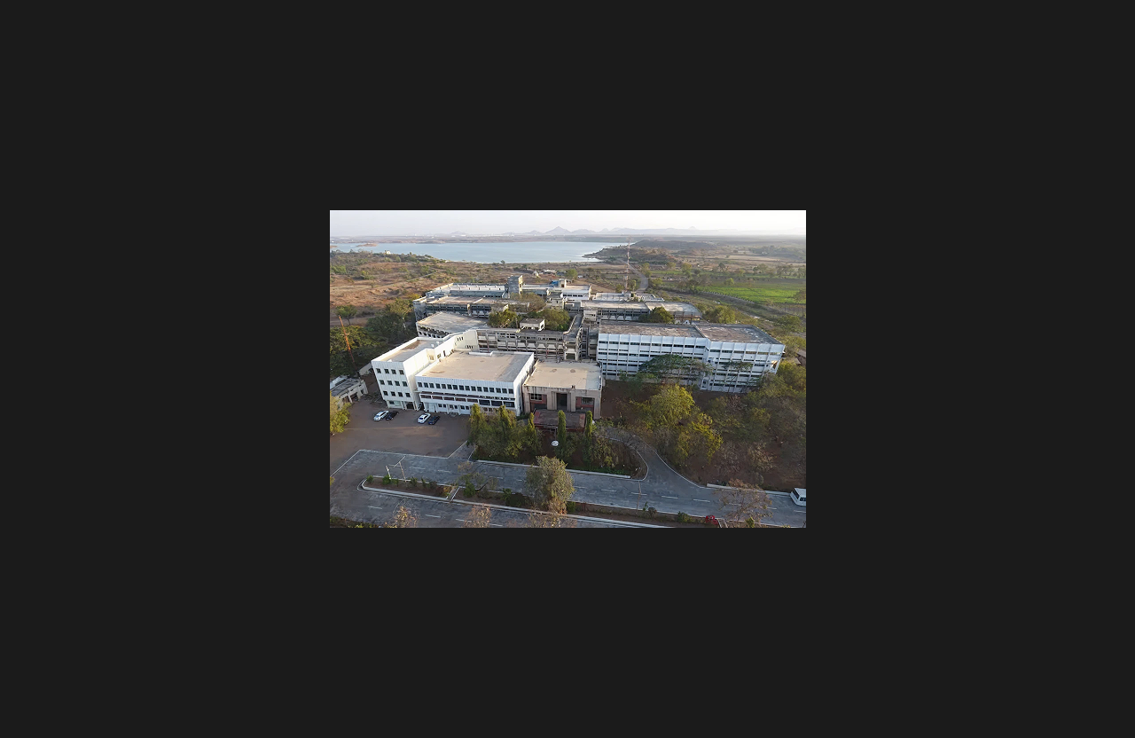 An aerial view of a large educational campus with multiple academic buildings, surrounding greenery, road access, and a water body visible in the background against a hilly landscape.