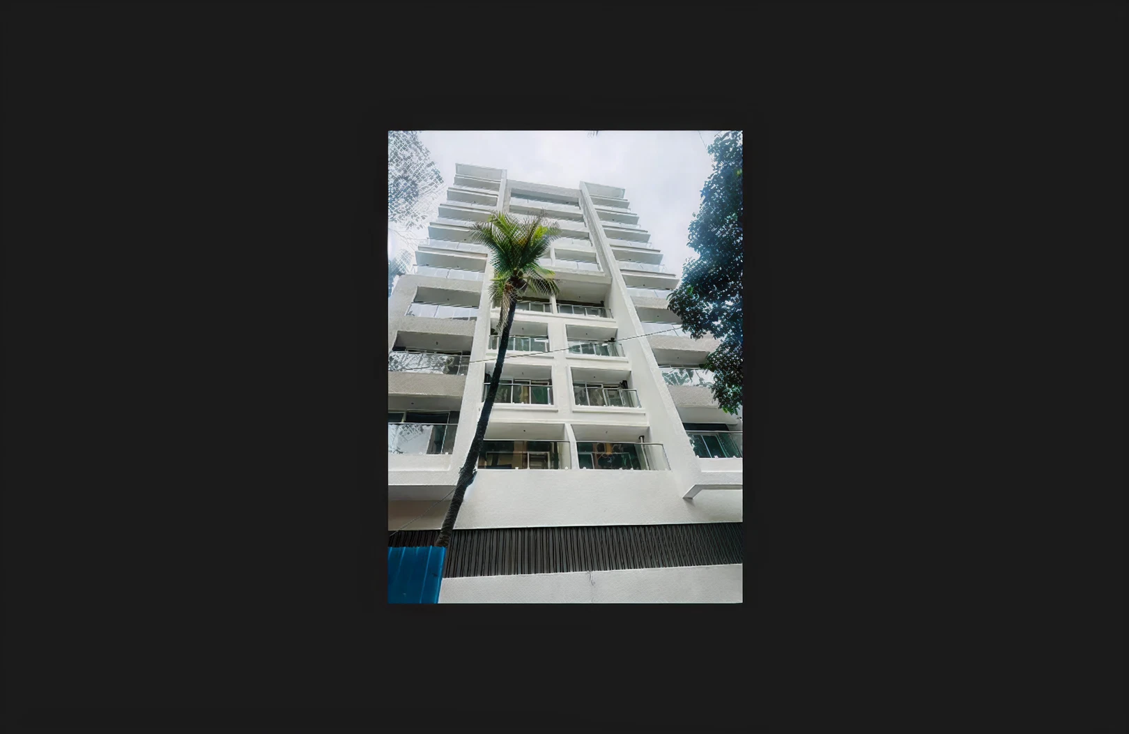 A modern white residential building viewed from a low angle, showing multiple balconies and clean geometric lines. A tall palm tree stands in the center foreground leaning slightly upward toward the facade, with other tree branches visible on both sides and a cloudy sky above.