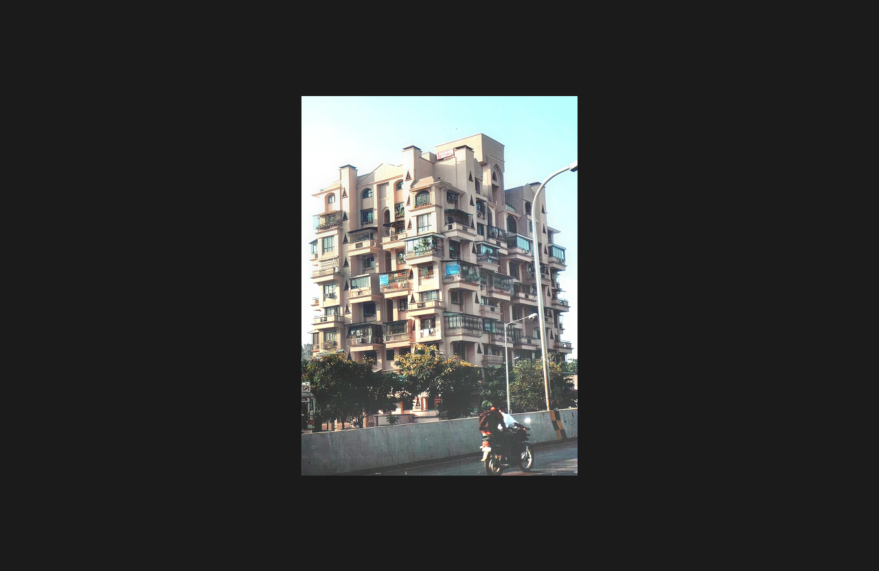 A multi-storey residential apartment building with a beige exterior and multiple projecting balconies featuring glass railings and awnings, photographed from a roadside view with trees, streetlights, and a motorbike passing in the foreground.
