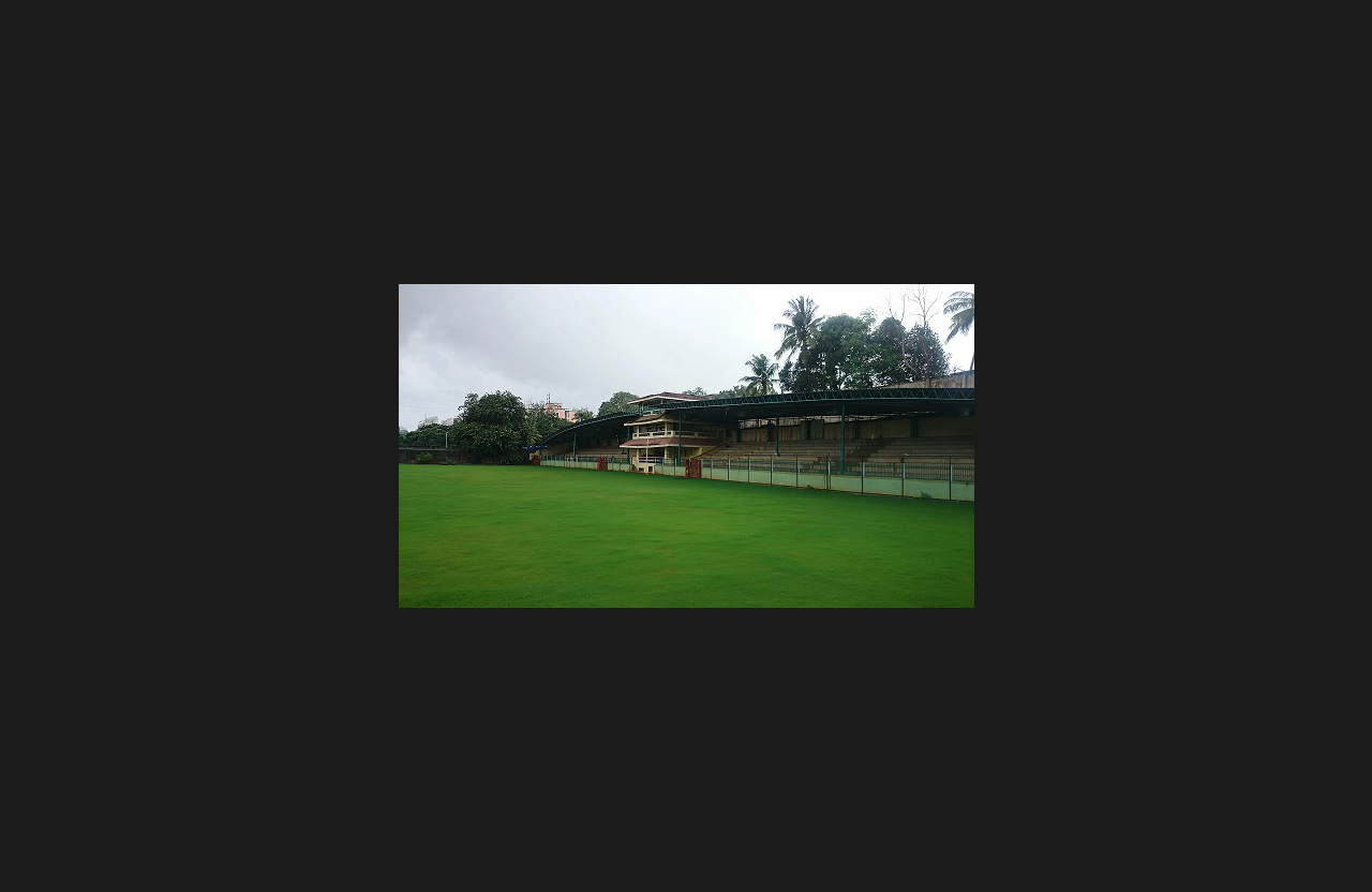 A wide open sports ground with a well-maintained grass field and covered spectator stands in the background, surrounded by trees and floodlight poles, under cloudy weather.