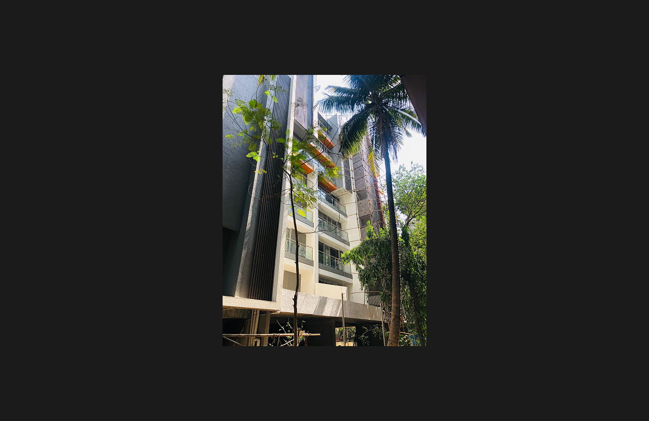 A tall premium residential building with glass balconies and vertical architectural fins, viewed through surrounding trees and a coconut palm, showcasing modern design and natural integration.