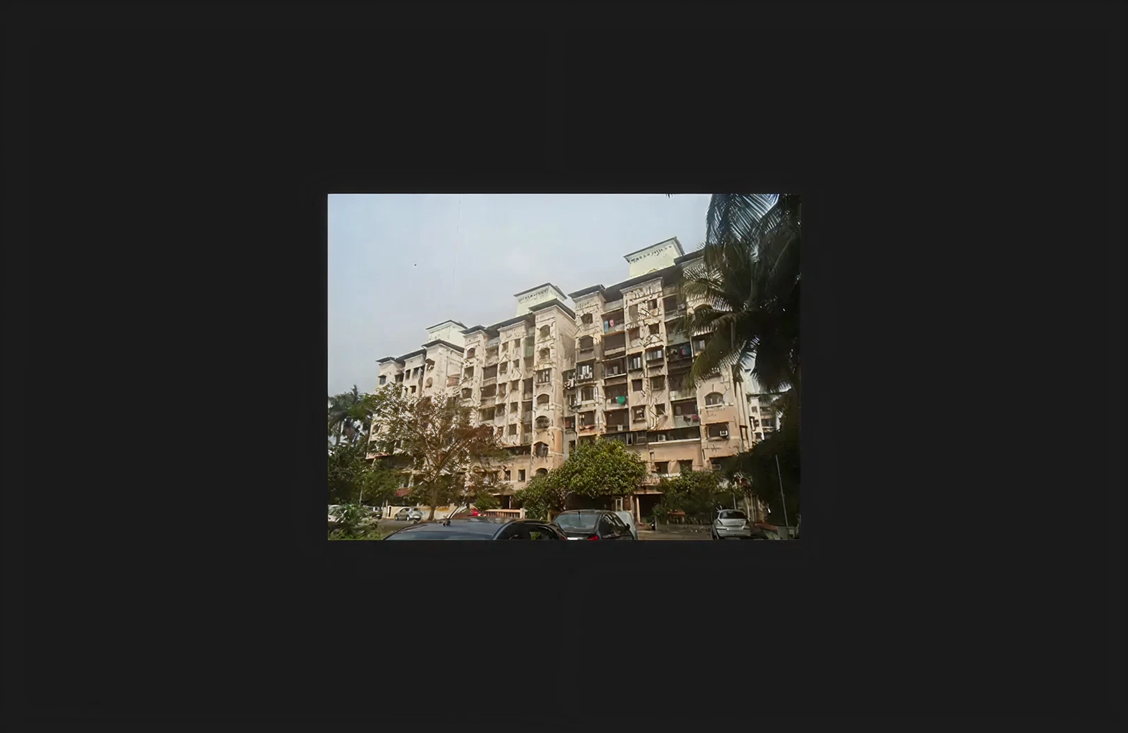 A large multi-wing residential housing complex showing an aged exterior prior to refurbishment, with visible façade wear, balcony structures, and uniform window arrangements. The building is surrounded by landscaped greenery, parked vehicles, and internal road access, representing a redevelopment or maintenance project within an established residential community.