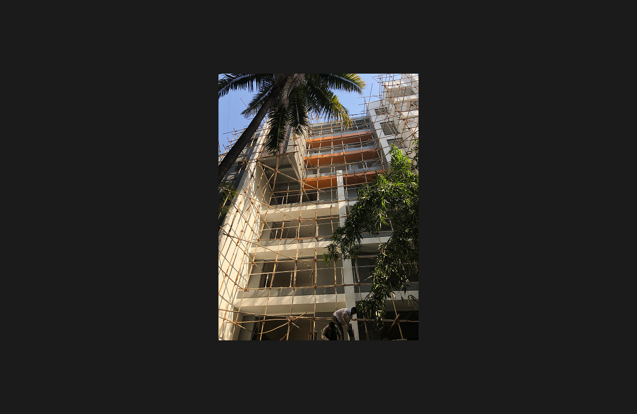 A mid-rise residential building under construction with bamboo scaffolding covering the front façade, surrounded by trees and a tall palm tree in the foreground, viewed from a low-angle perspective under clear daylight.