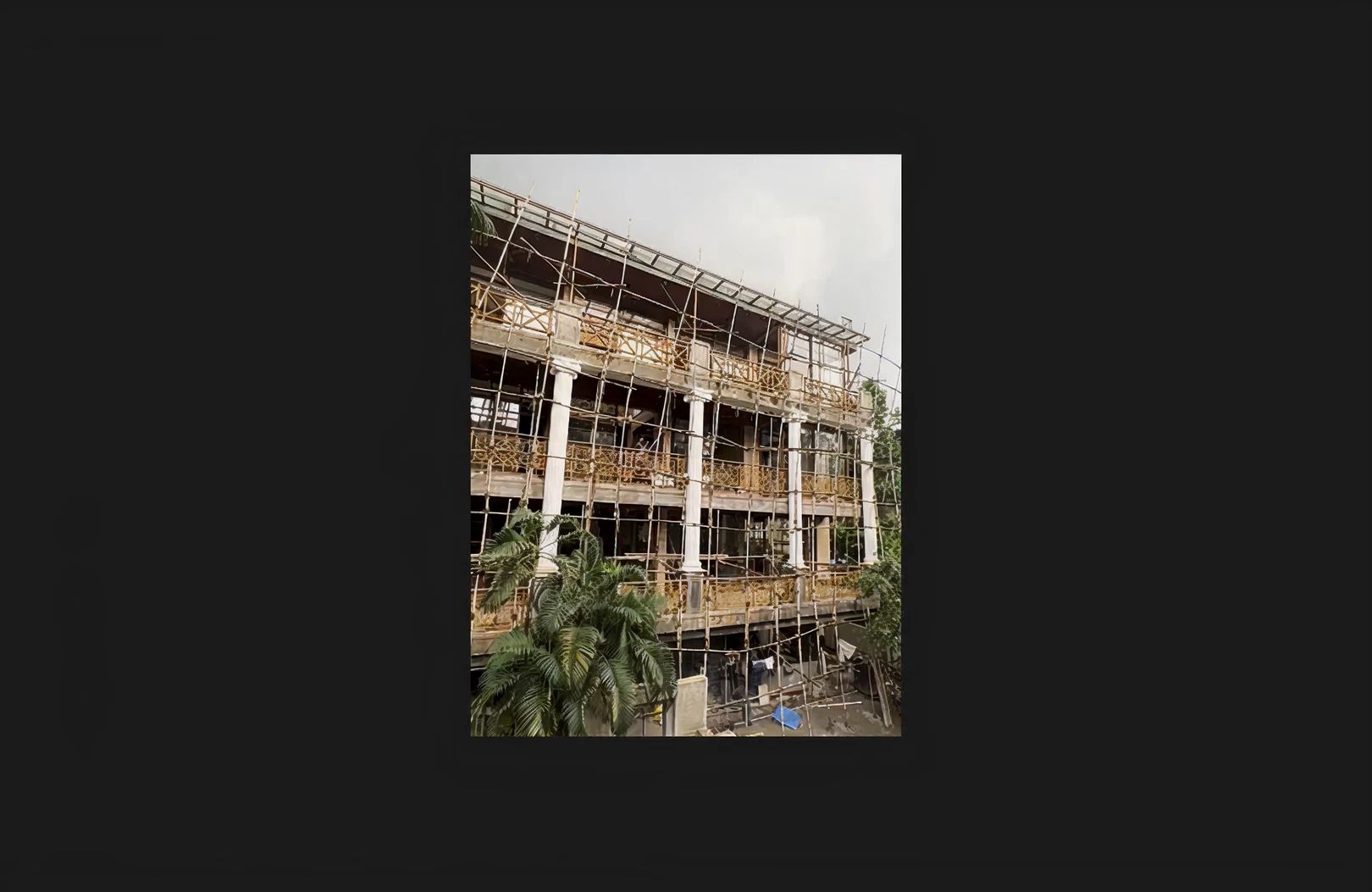 A multi-storey building under construction with full external scaffolding in place, showcasing structural framework, elevation detailing, and classical column elements. The site reflects active construction progress with safety structures installed and finishing work underway on the façade, surrounded by landscaping and greenery.