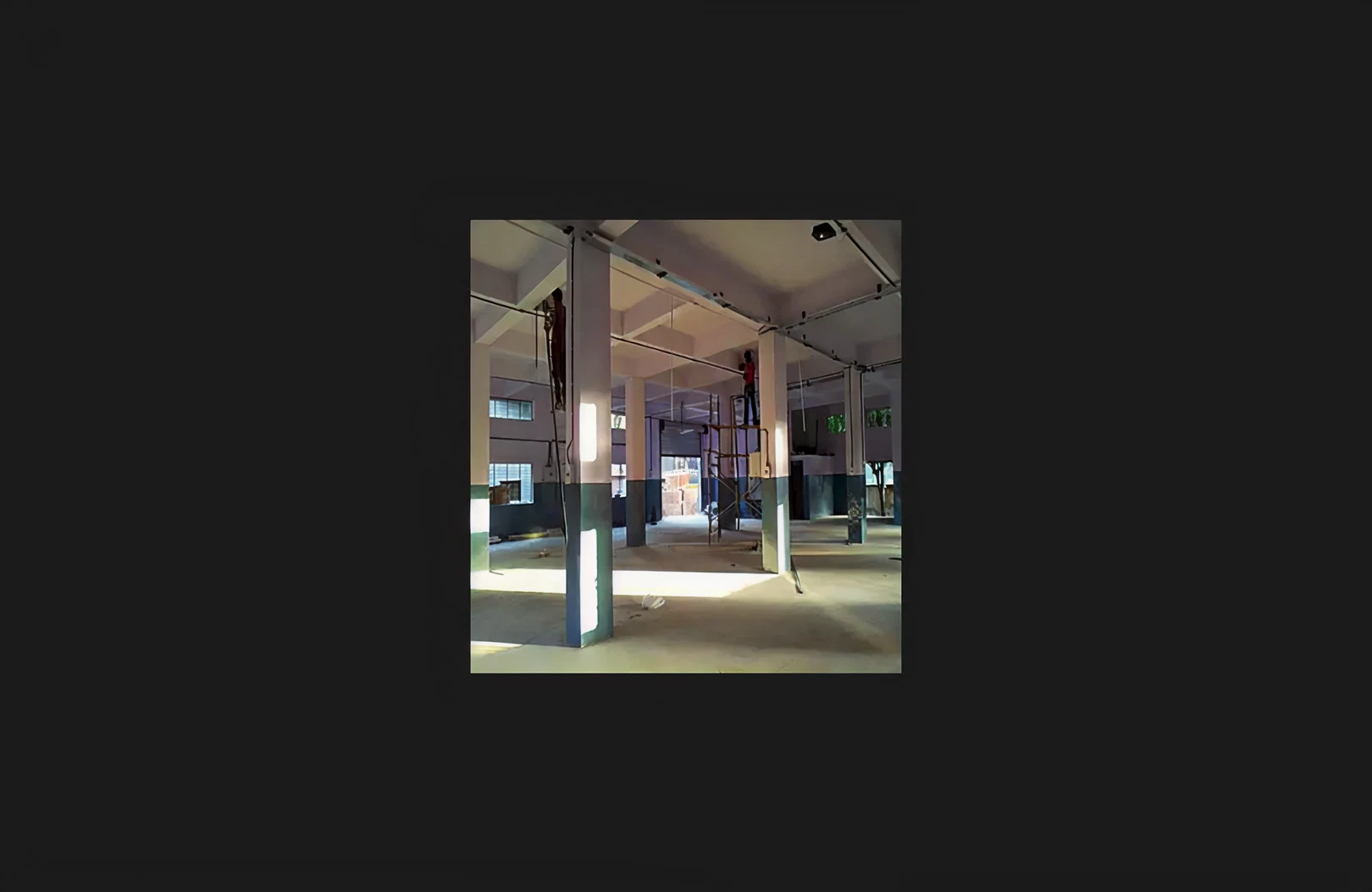 An active interior construction site showing structural columns, scaffolding, and workers carrying out ceiling and electrical installation work. The space features an open floor layout with reinforced concrete columns and natural light filtering through large windows, indicating ongoing renovation and interior development progress.