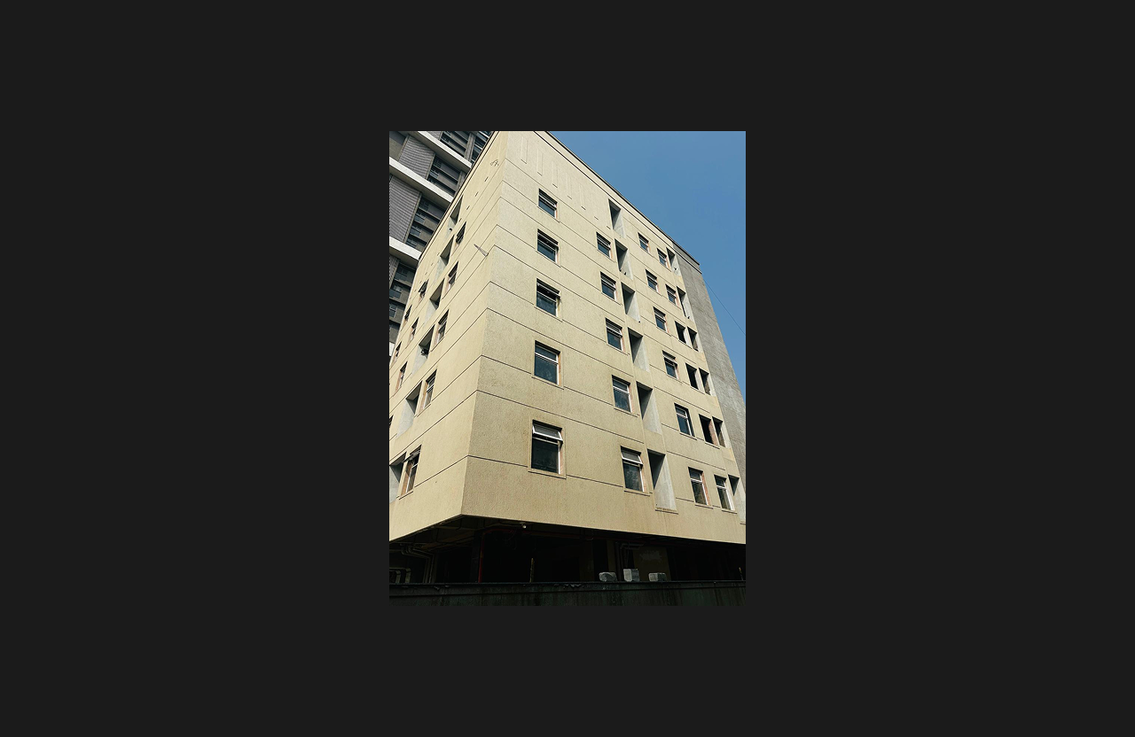A mid-rise institutional or commercial building with a beige textured exterior façade and evenly aligned rectangular windows, photographed from a low-angle perspective against a clear blue sky.