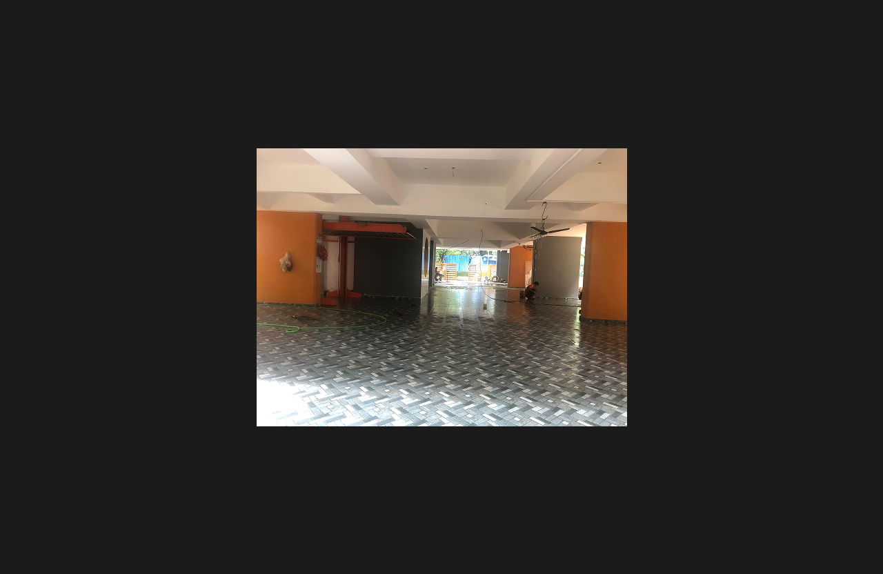 An interior ground-level space of a building under finishing stage, featuring newly installed patterned tile flooring, exposed ceiling structure, workers cleaning and completing final surface work, and open access to exterior areas.