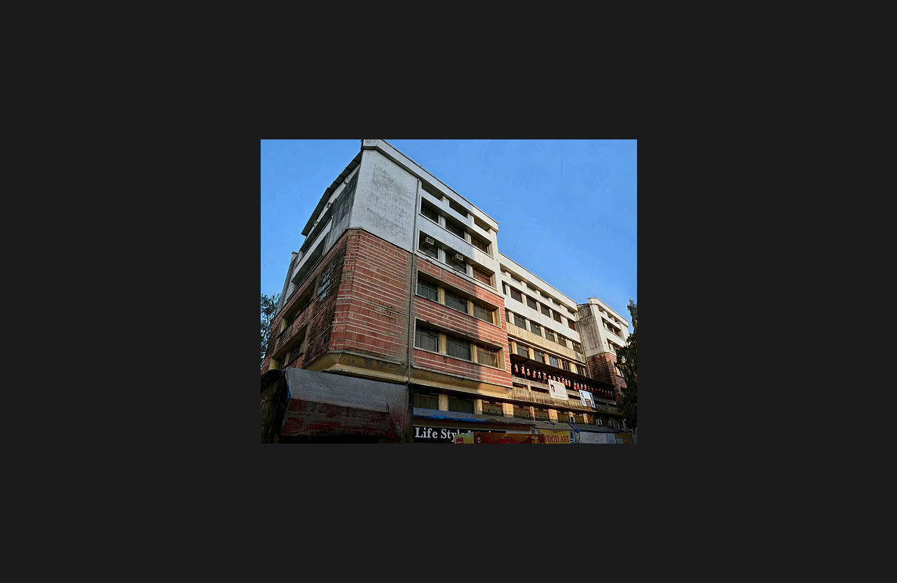 A multi-storey commercial and residential building with an older exterior featuring weathered white and red brick-textured façade, retail shops at the ground level, and multiple balconies above, captured from a street-level low-angle view under a clear blue sky.