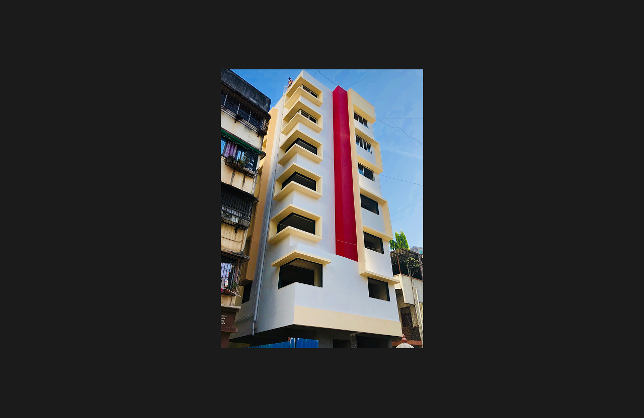 A newly completed mid-rise residential building with a clean cream and white façade and a bold vertical red accent stripe, featuring geometric window projections and a stilt-level entrance, photographed from a low-angle perspective.