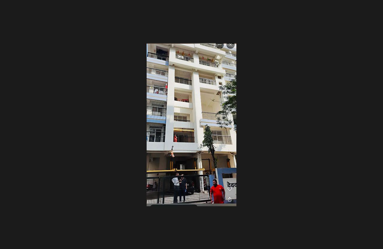 A completed residential building with a modern white façade and black railing balconies, viewed from the ground level near the entrance gate, with residents and workers standing by the gate and a small tree in the foreground.