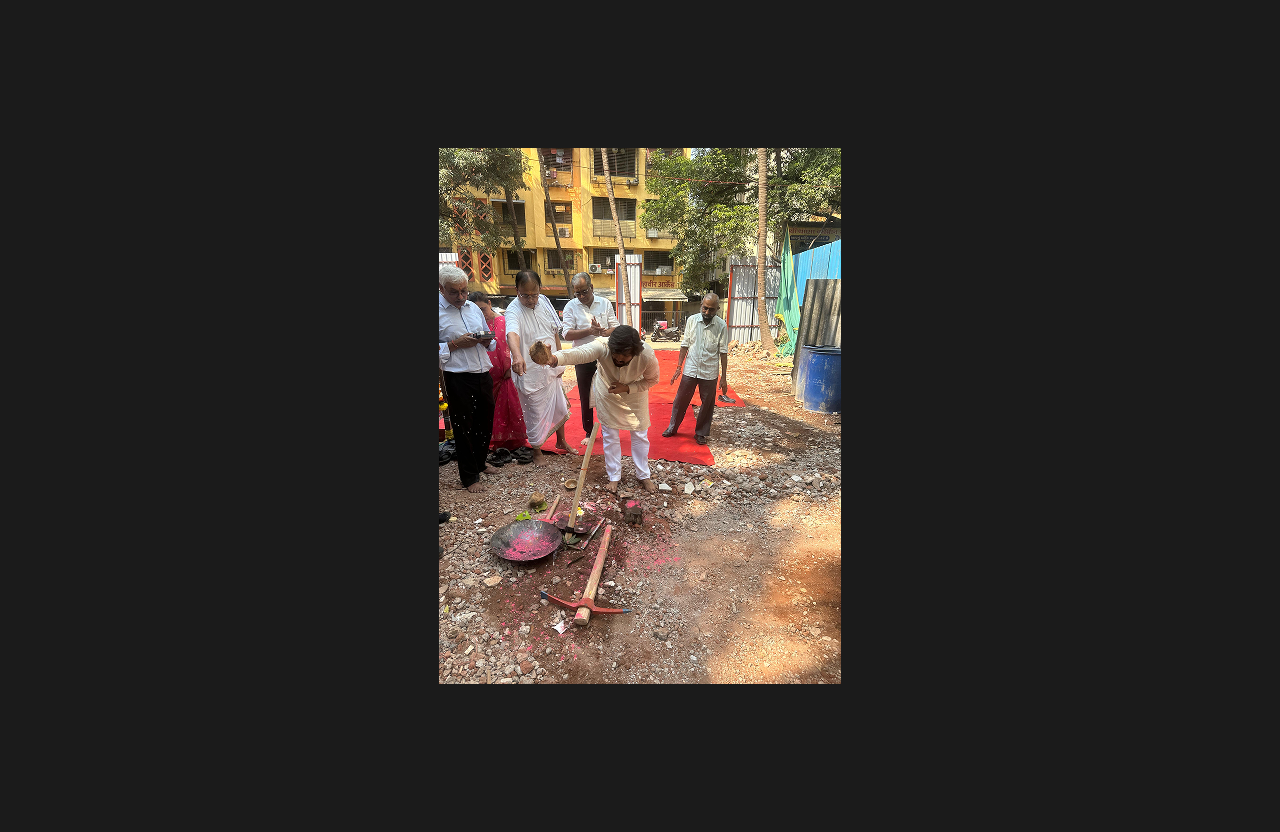 A traditional groundbreaking ceremony at a construction site, with a man performing ritual prayers using a tool and ceremonial offerings, surrounded by attendees and a red carpet walkway.