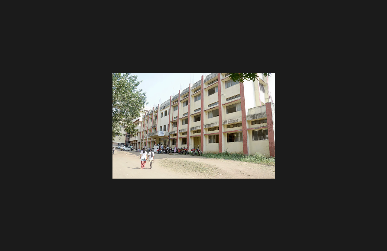 A multi-storey educational building with a cream-colored exterior and vertical red structural elements, featuring open corridors and students walking near the entrance, with motorcycles parked along the front.