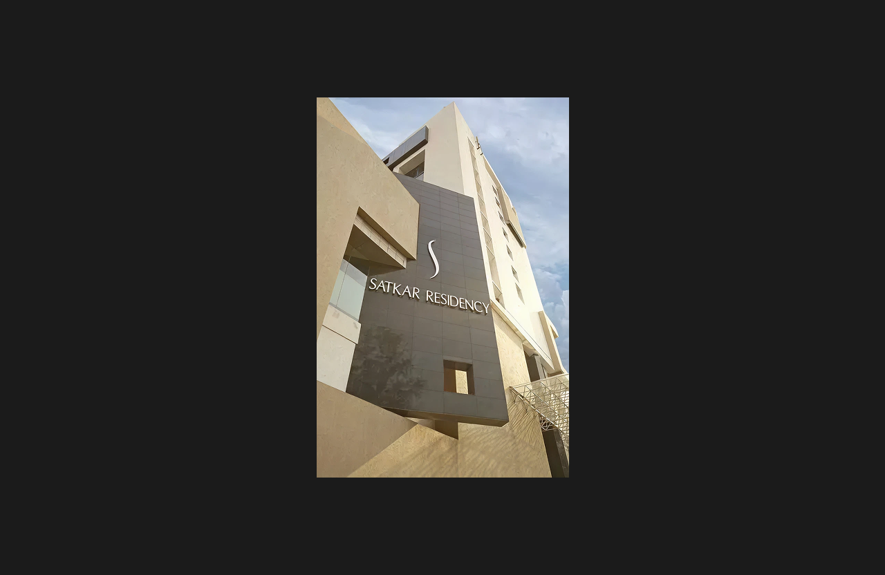 A modern high-rise building facade featuring sleek beige and grey cladding with the name ‘Satkar Residency’ prominently displayed on a vertical architectural panel, photographed from a low-angle perspective against a cloudy sky.