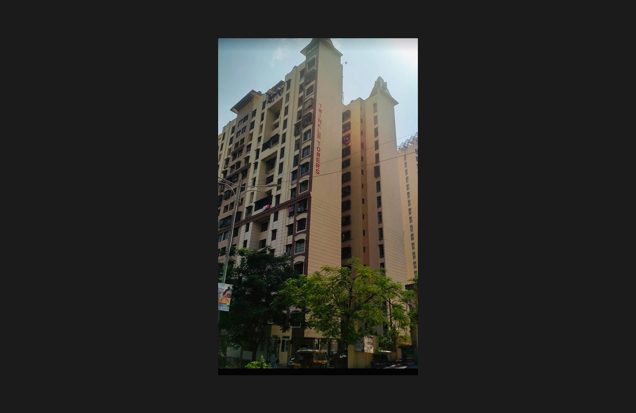 A high-rise residential building with vertical beige exterior finishes and the name ‘Triveni Towers’ displayed prominently along the side elevation, photographed from street level with surrounding trees and urban activity.