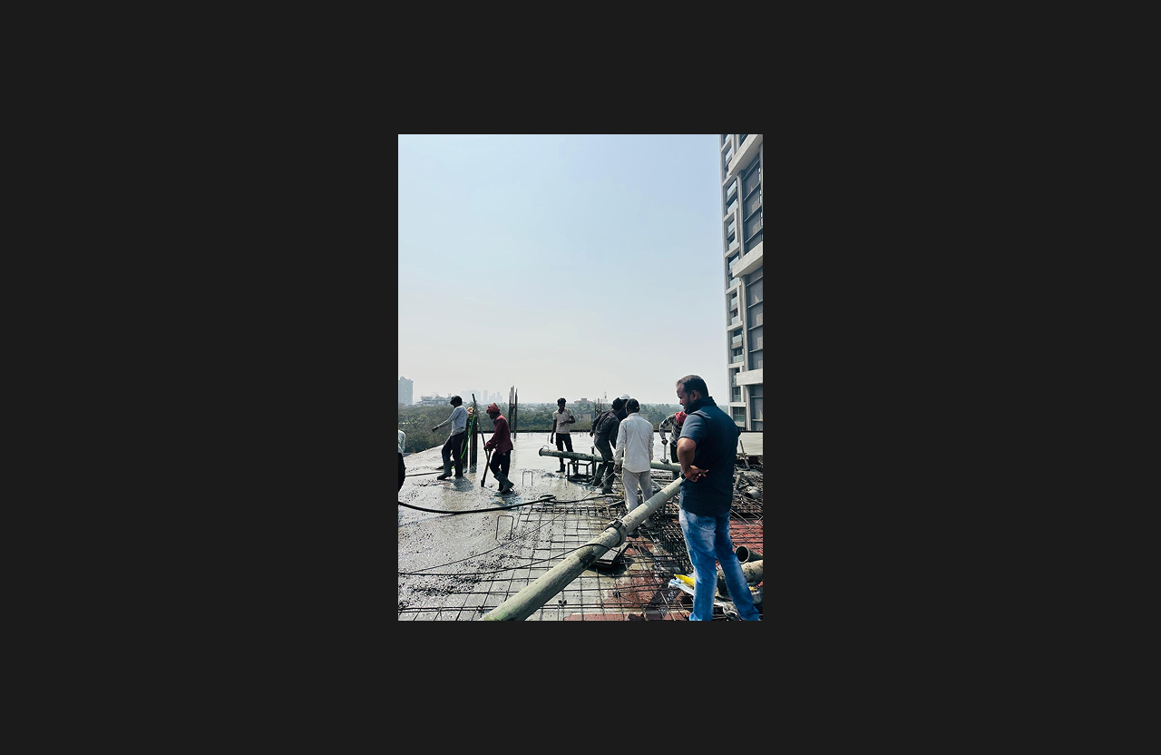 Construction workers carrying out concrete slab casting on the upper floor of a building, using pumping equipment and reinforcement bars visible in the foreground, with a site supervisor observing and an urban skyline in the background under clear daylight.