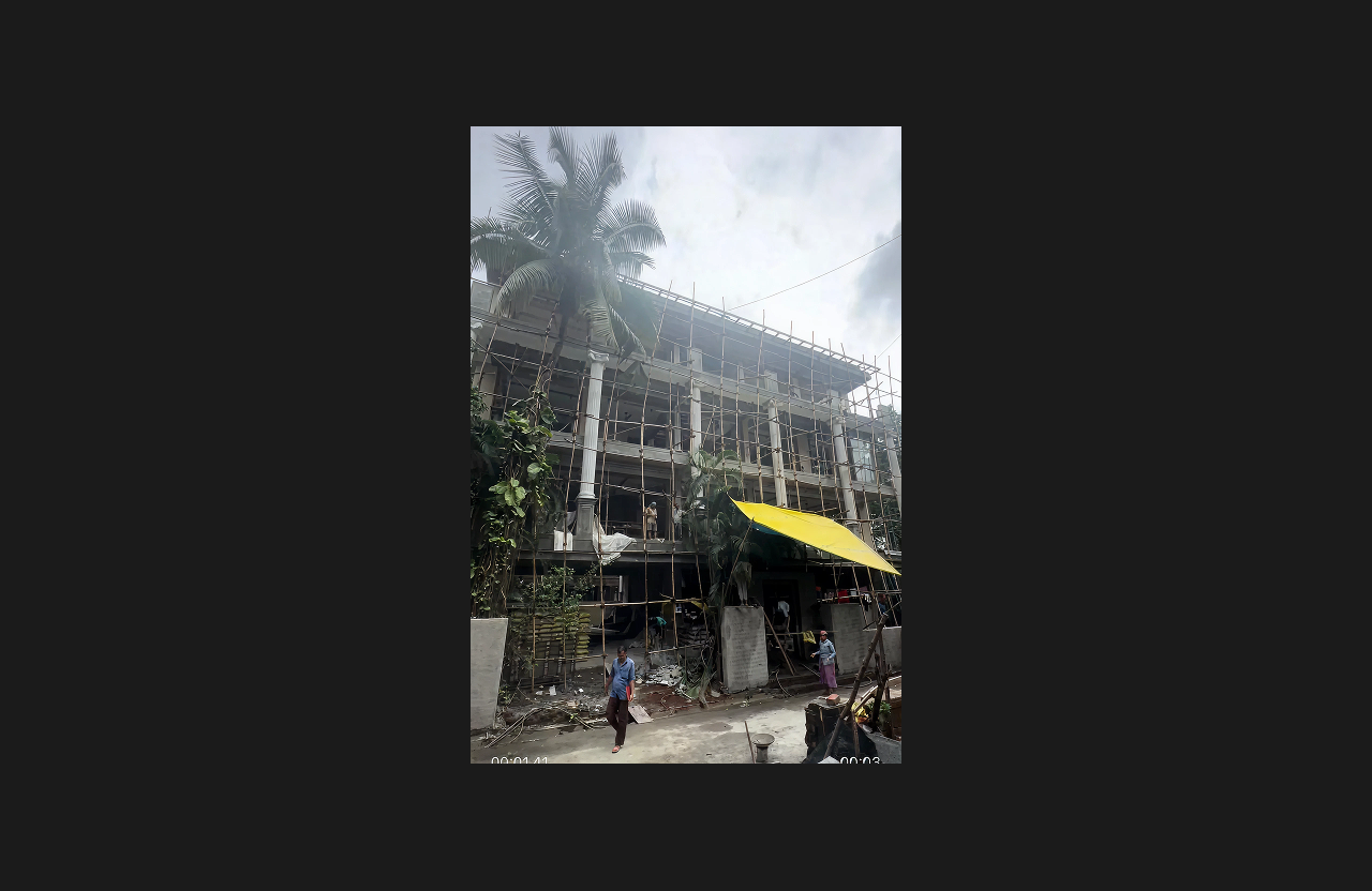 A renovation project in progress with extensive scaffolding around a multi-storey building, construction workers and materials at the ground level, and trees and a temporary covered shed near the entrance.