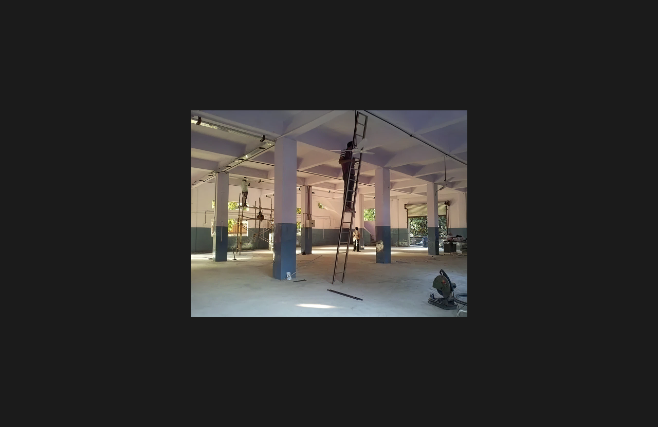 A large open interior space under renovation, with a worker on a tall ladder fixing ceiling utilities, tools and construction materials on the floor, and multiple support columns visible throughout the hall.