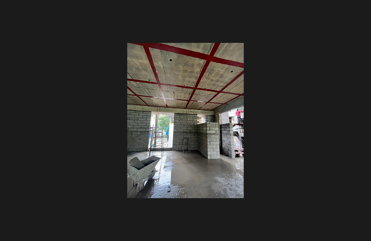 An under-construction interior space with wet concrete flooring, exposed brick walls, and a ceiling grid marked with red structural layout lines, showing construction tools, scaffolding, and natural light entering through open wall sections.