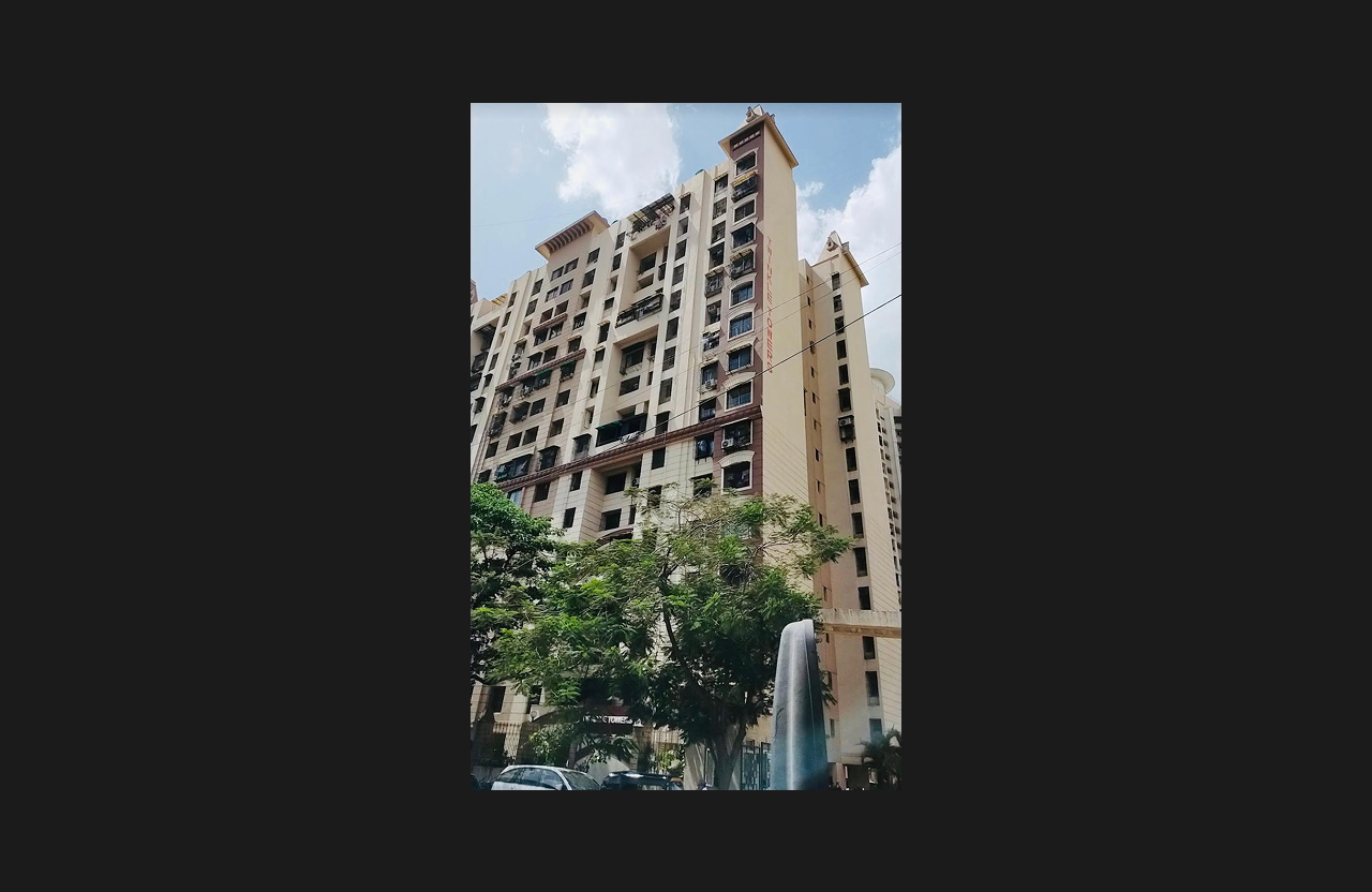 A tall residential apartment tower with beige façade and arched balcony openings, captured from a ground-level perspective with trees and greenery in the foreground under a partly cloudy sky.