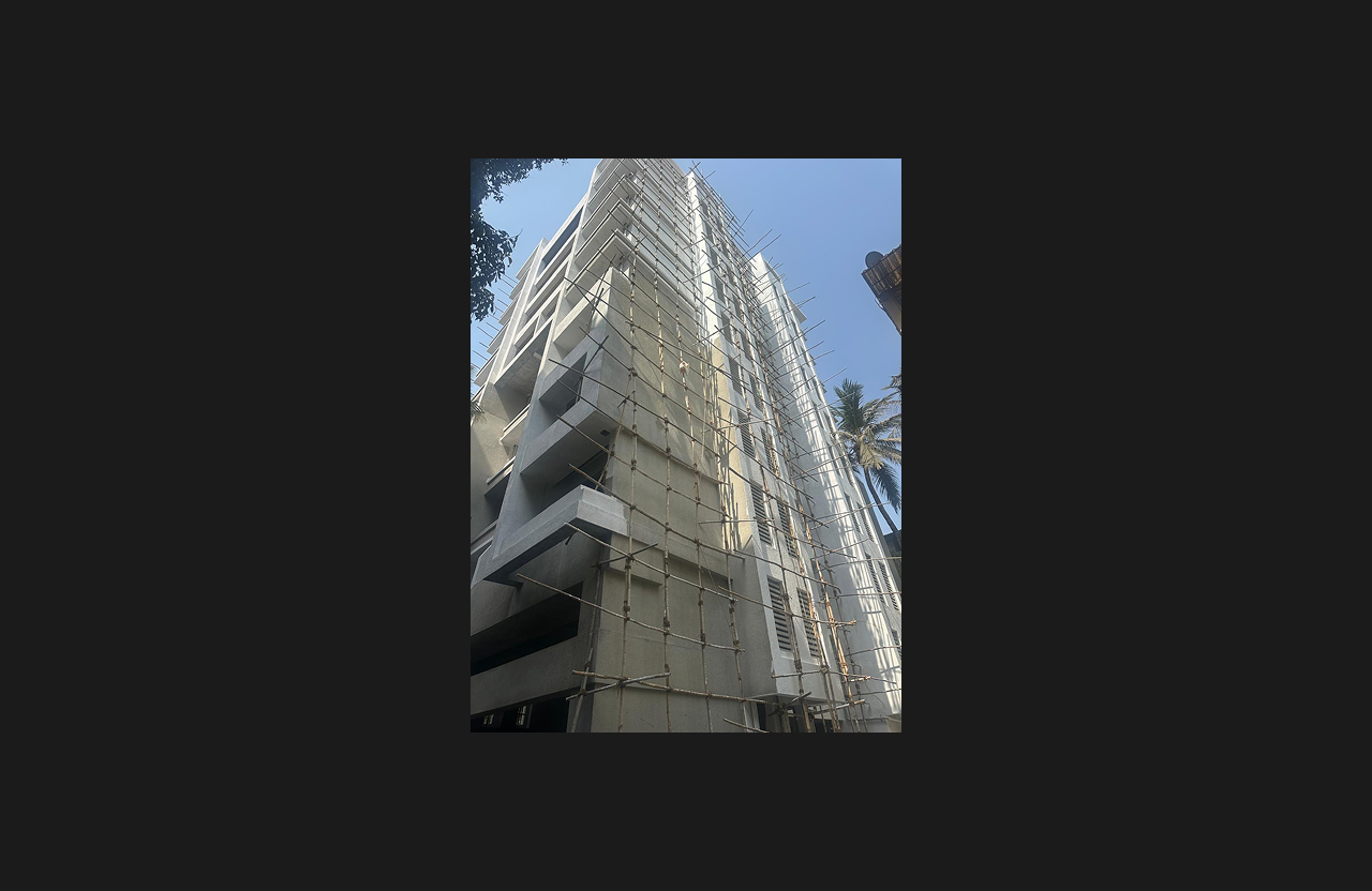 A high-rise residential building under construction with full external scaffolding surrounding the façade, featuring white and grey elevation elements, photographed from a street-level perspective.