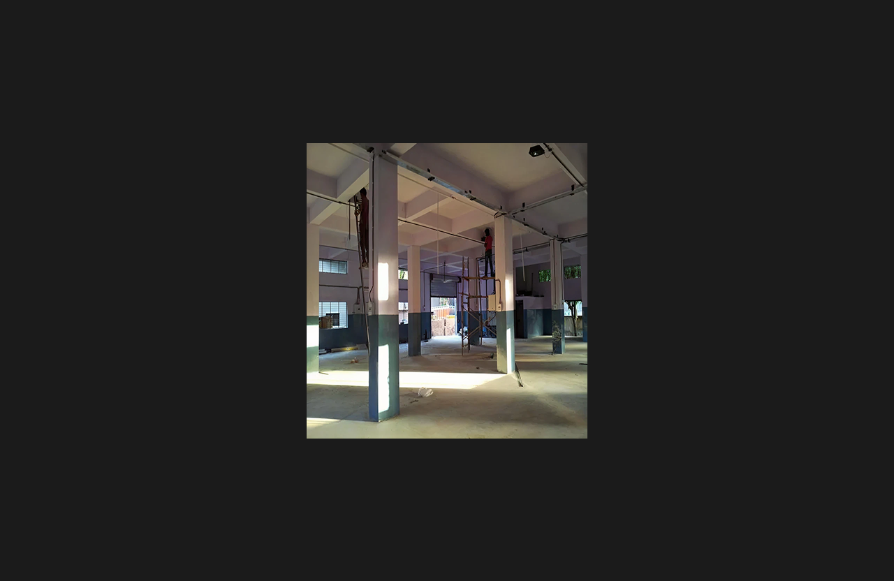 Interior construction site showing workers installing electrical and ceiling infrastructure on scaffolding, with exposed columns, unfinished flooring, and natural light entering through surrounding windows.