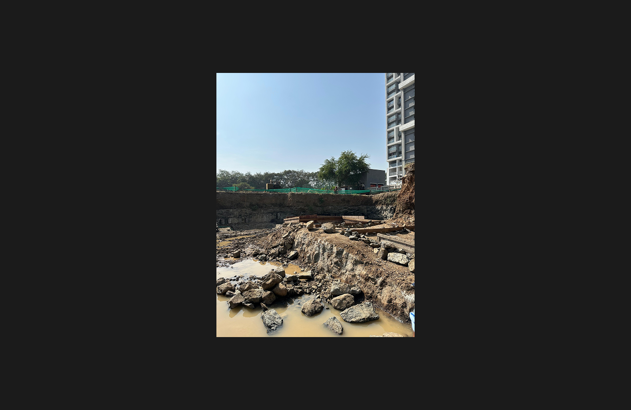 Excavation site with exposed soil layers, scattered construction rocks, and water accumulation in the work area, with foundation preparation in progress and a nearby residential building and trees visible in the background under a clear sky.