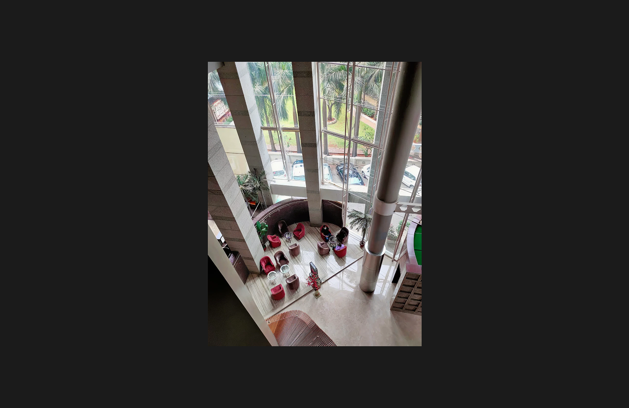 A luxurious double-height lobby space with floor-to-ceiling glass windows, polished marble flooring, modern seating with red lounge chairs, indoor plants, and tall structural columns offering views of the outdoor greenery.