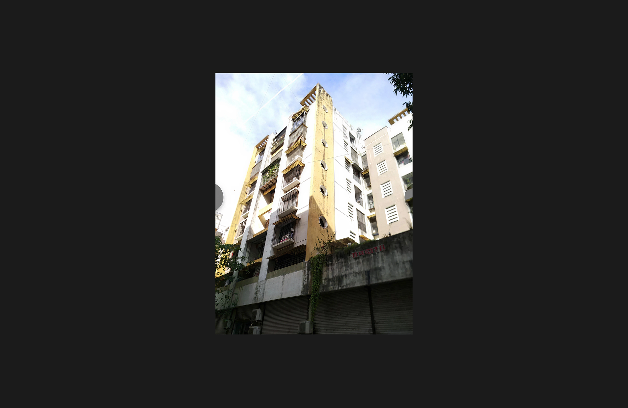 A mid-rise residential apartment building with a white and yellow exterior, vertical architectural fins, and circular window details, photographed from a low-angle street view with closed commercial shutters at ground level and greenery climbing the facade.