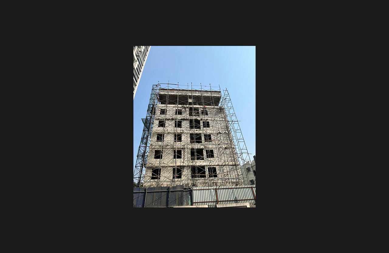 A multi-storey building covered entirely in external scaffolding during construction work, with open window frames and unfinished façade visible, captured from a low-angle perspective under bright daylight.