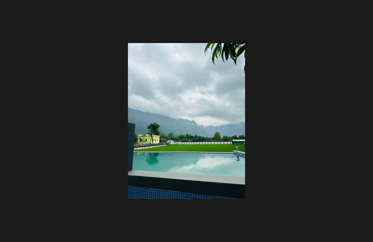 A still-water swimming pool reflecting cloudy skies, overlooking a lush green lawn and distant mountain range, creating a serene natural retreat setting.