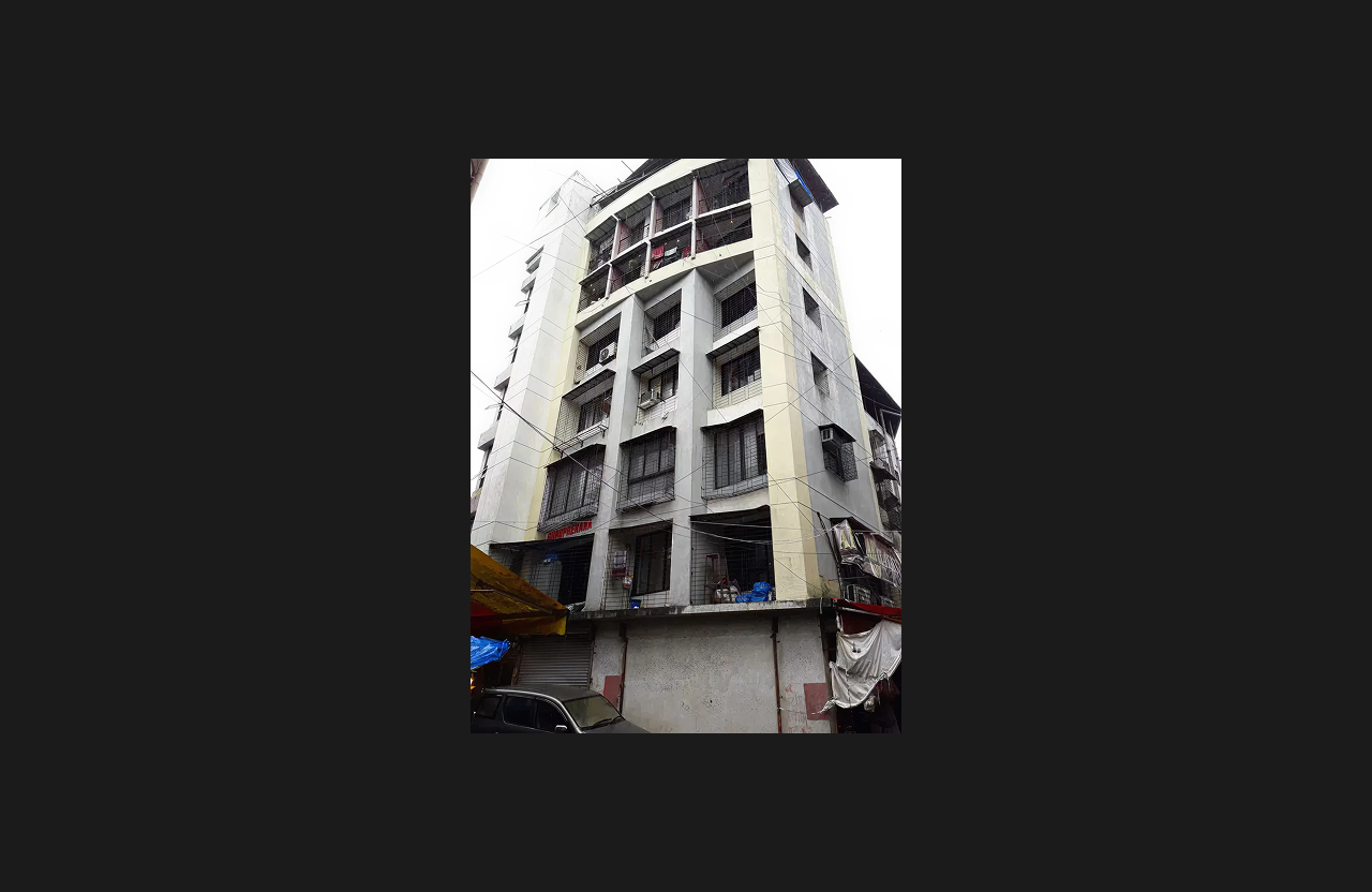 A multi-storey residential building with a cream and white exterior, grid-patterned balcony grills, and commercial shutters at the ground level, photographed from a low-angle street view with surrounding market structures and vehicles.
