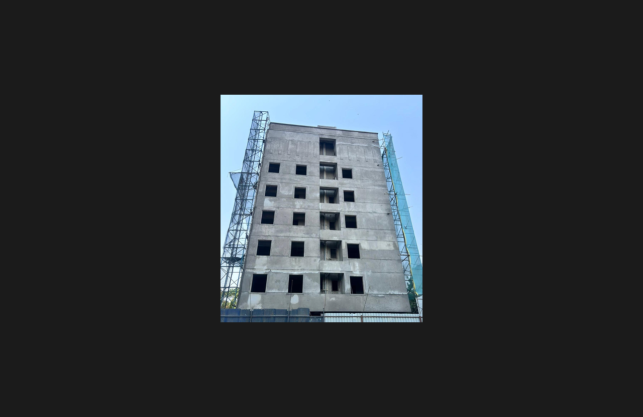 A mid-rise building under construction with completed concrete structure and window openings, supported by metal scaffolding on both sides, photographed from ground level against a clear sky.
